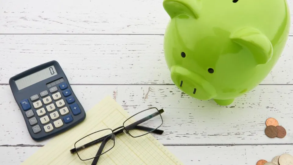 A green piggy bank next to a calculator, reading glasses and a few coins
